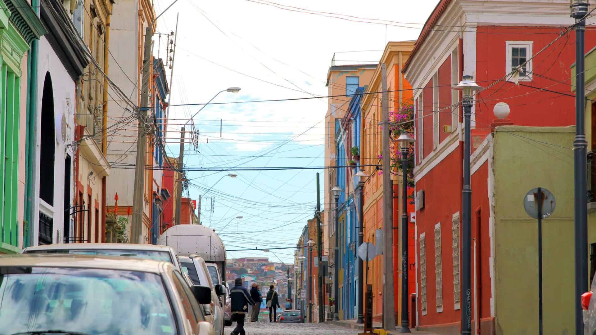 Colorful buildings line a cobblestone street with cars parked and people walking under a sky crisscrossed with wires.
