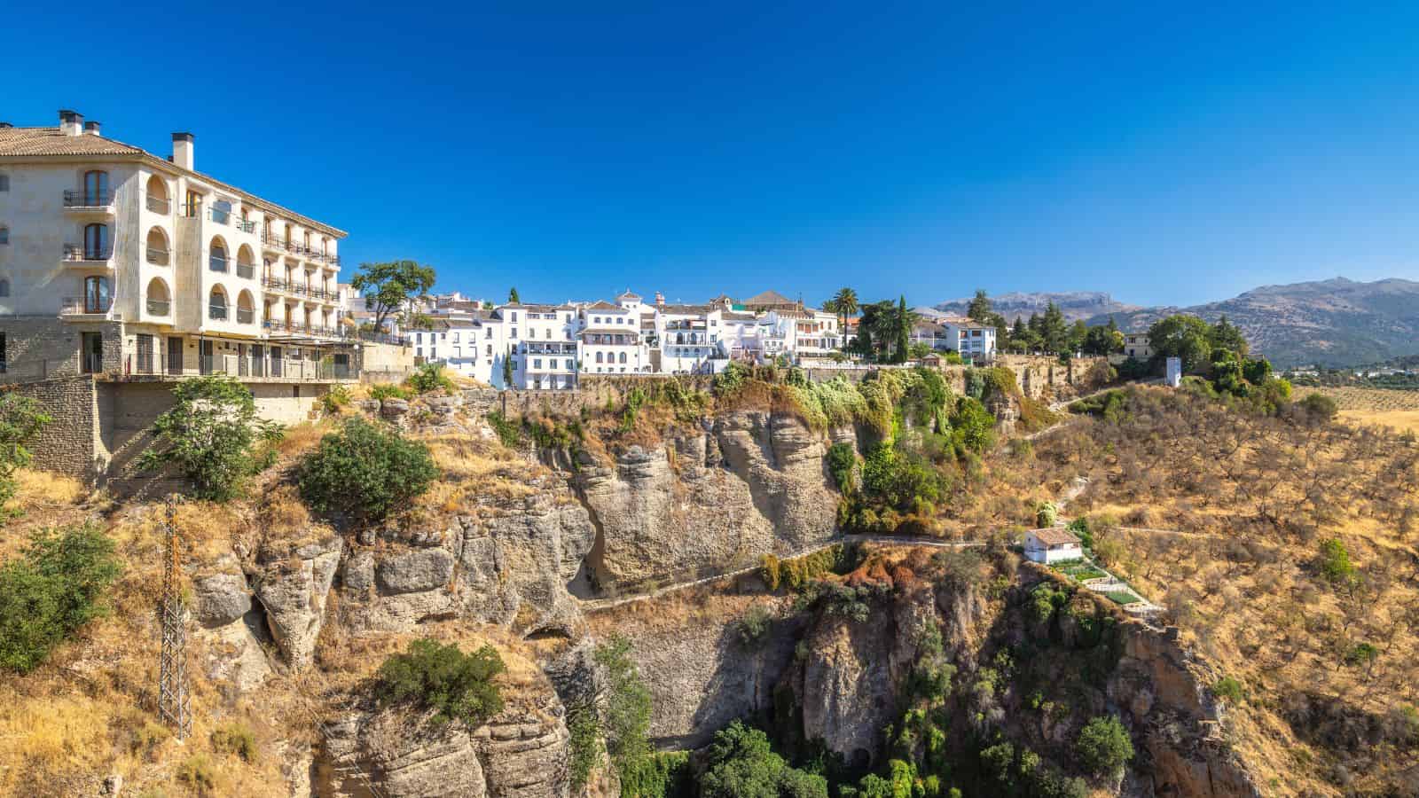 White buildings perched on a cliff above a deep gorge in a sunny, mountainous landscape.