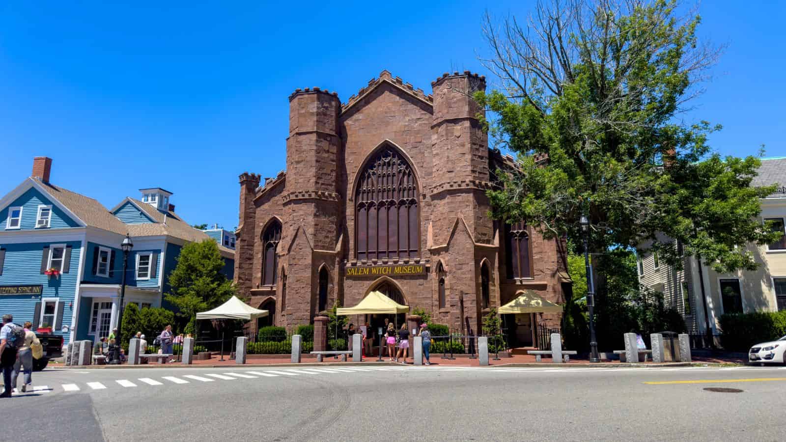 Brown Gothic-style Salem Witch Museum with large arched windows, flanked by blue and gray houses and trees.