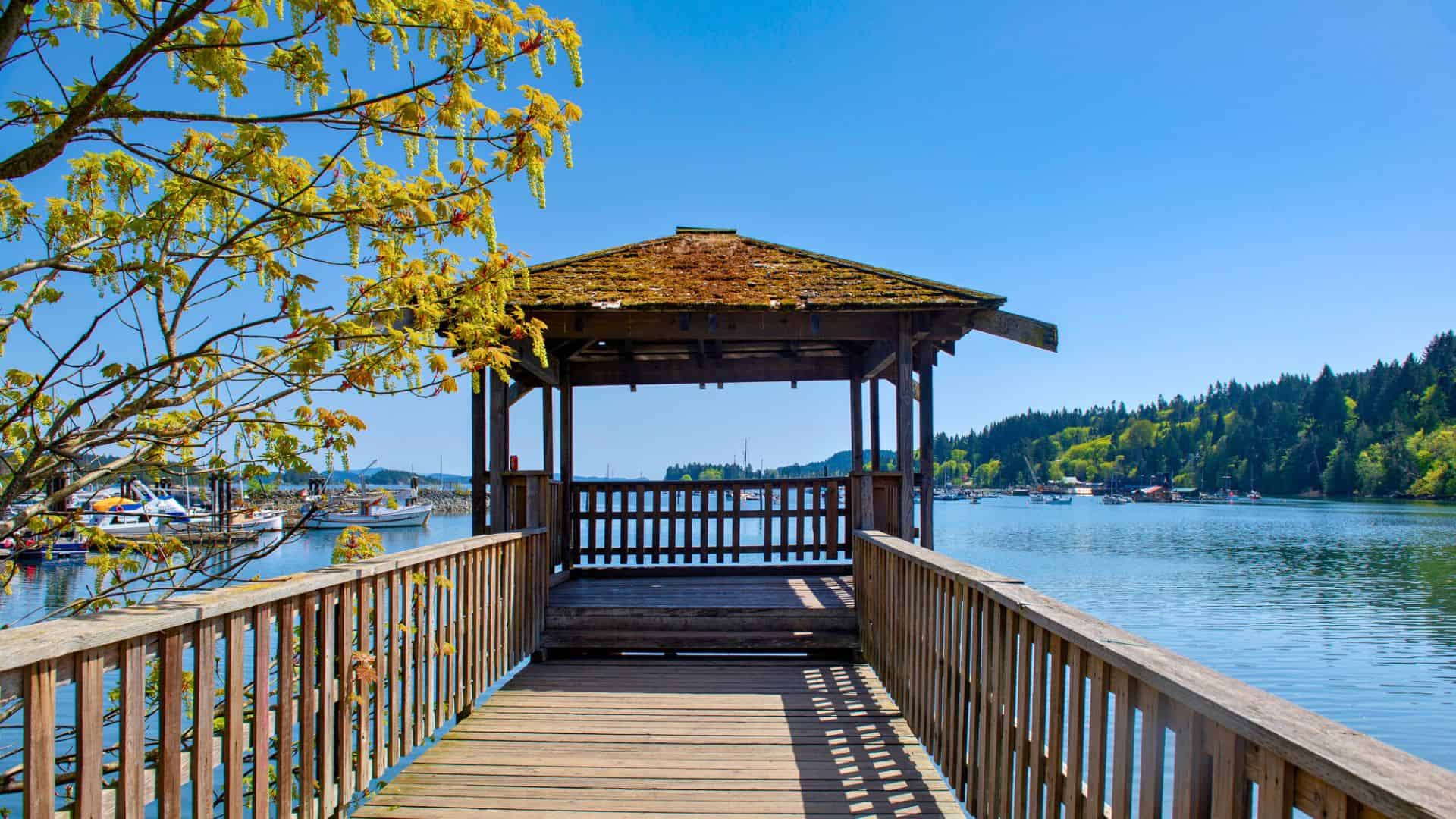 A wooden gazebo on a pier overlooks a calm lake, surrounded by trees under a clear blue sky.