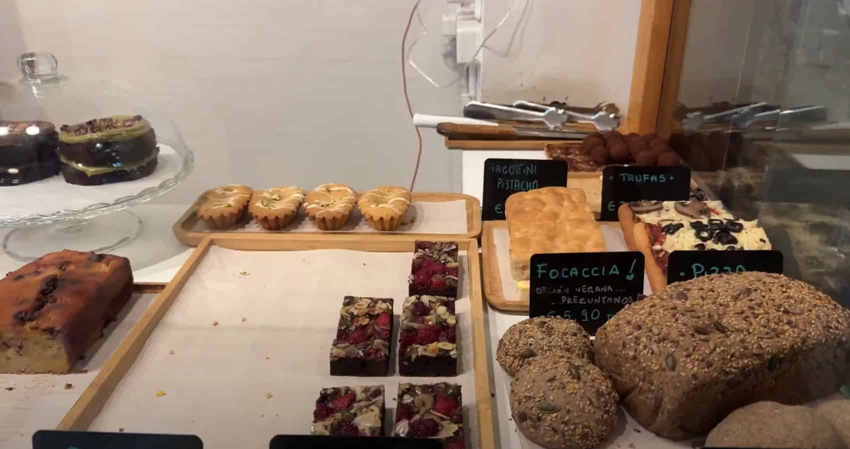 A bakery display with assorted breads, pastries, cakes, and handwritten signs on a wooden counter evokes the vibrant charm of Barcelona markets, known for their fresh food and artisanal treats.