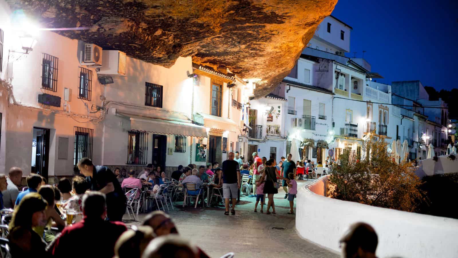 People dine outdoors under a large rock overhang in a charming, well-lit village at dusk.