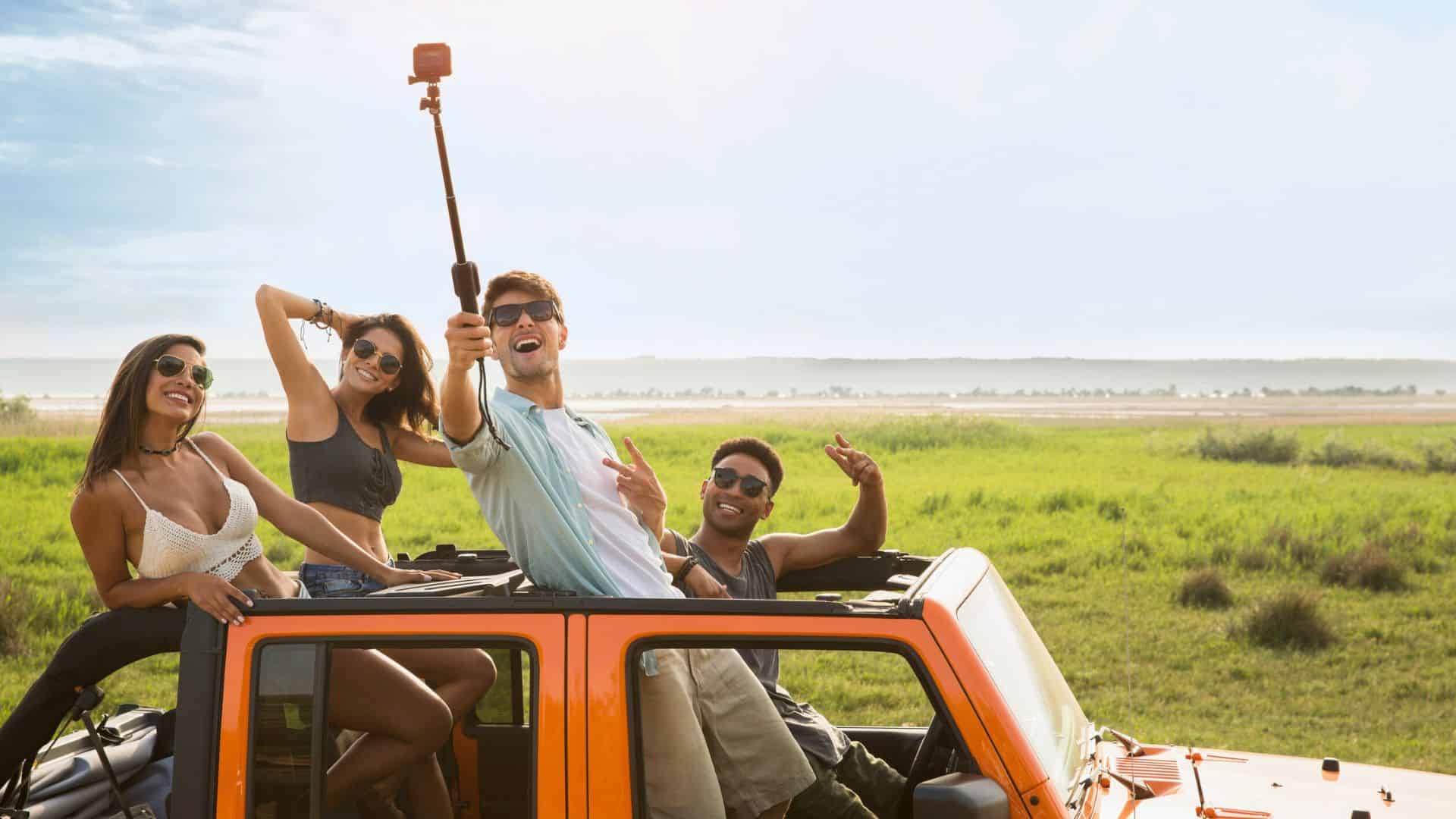 Four friends in a jeep taking a selfie with a selfie stick in a grassy outdoor landscape on a sunny day.