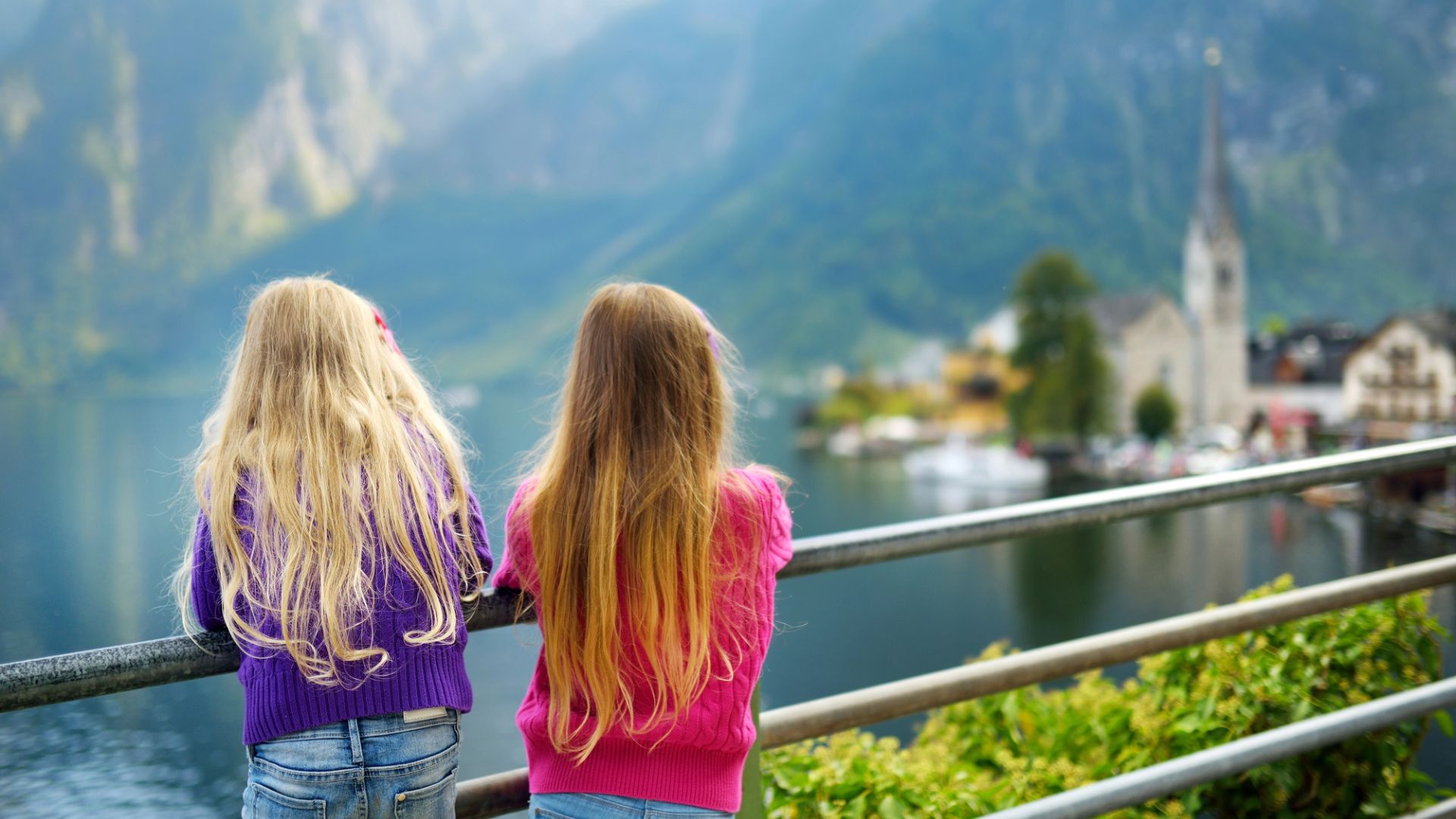 Two young girls with long hair look at a lake and a distant village with mountains in the background.