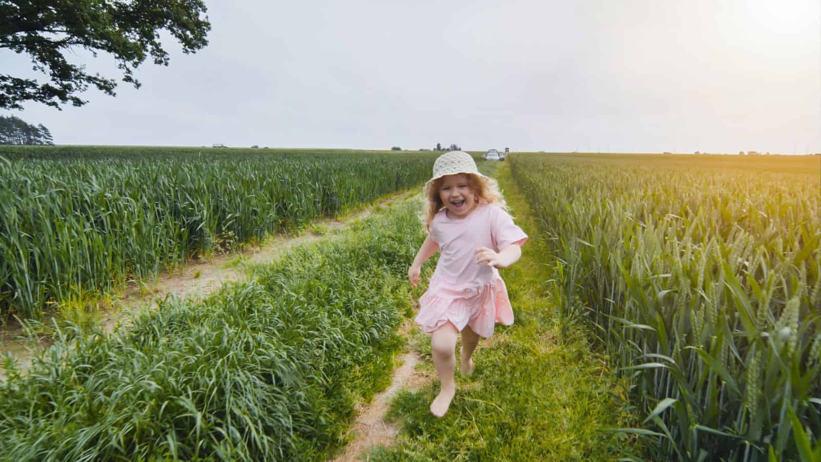 Smiling girl in a pink dress runs barefoot along a grassy path between green fields on a sunny day.