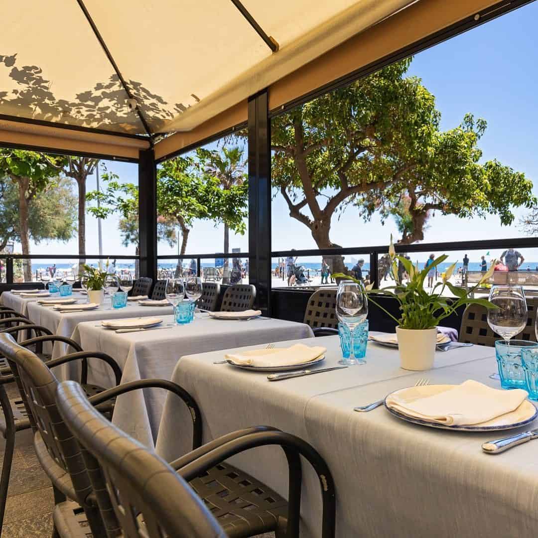 Chic outdoor restaurant with neatly set tables under a canopy, overlooking trees and Barcelona’s sunny beachfront promenade—perfect for lovers of beachfront restaurants.