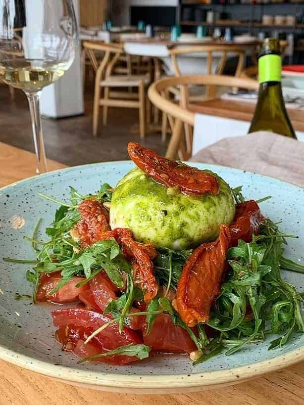 A plate of burrata cheese on arugula, tomatoes, and sun-dried tomatoes, next to a glass of white wine—perfect for enjoying at one of Barcelona’s best restaurants or chic beachfront restaurants.