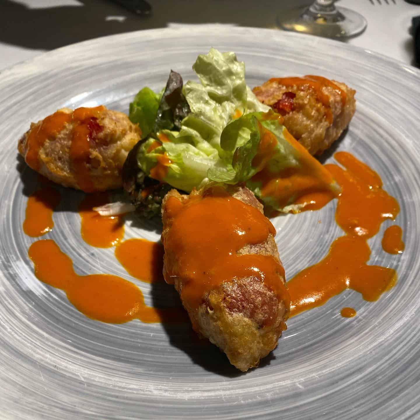 Three breaded croquettes with orange sauce and a small green salad on a gray plate, inspired by the fresh flavors found in Barcelona’s vibrant beachfront restaurants.