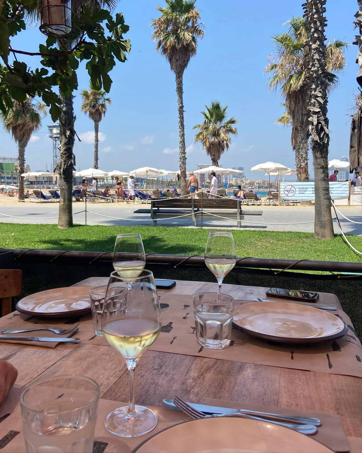 Table set for lunch with wine glasses by a beach, palm trees, and people sunbathing in the background—capturing the vibrant atmosphere of Barcelona dining at one of the finest beachfront restaurants Barcelona has to offer.