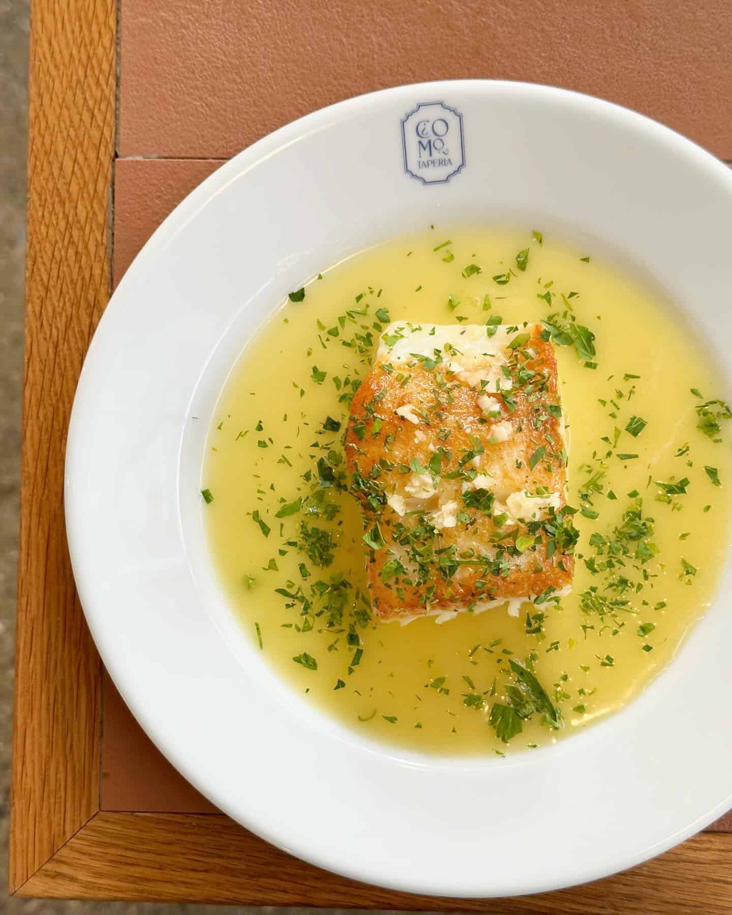 A piece of fish in yellow sauce, garnished with chopped herbs, on a white plate—reminiscent of dishes served at Barcelona’s vibrant beachfront restaurants.
