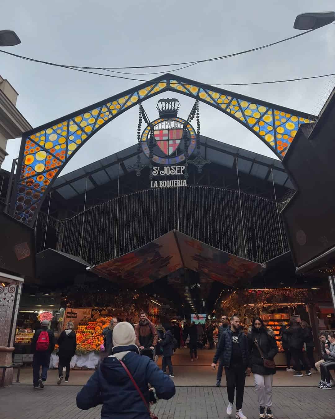 People walk outside the entrance to La Boqueria market in Barcelona, a must-visit among Barcelona markets, with colorful stained glass above.