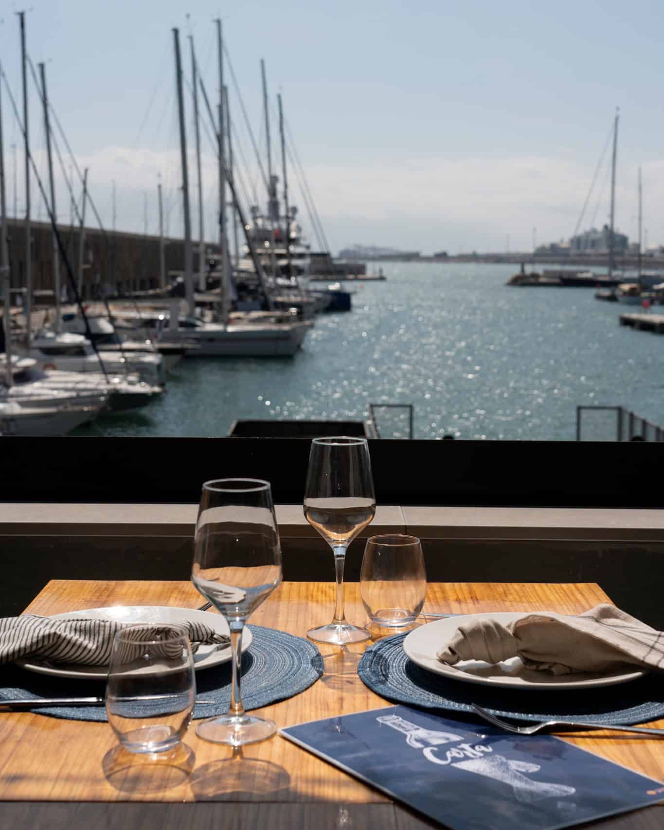 A table set for two overlooks a marina with sailboats on a sunny day, capturing the charm of Barcelona restaurants with a view.