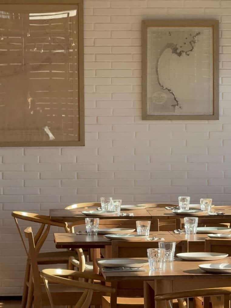 Sunlit wooden dining tables with plates and glasses, set against a white brick wall with framed art—perfect for those seeking the charm of Barcelona dining found in the best restaurants Barcelona has to offer.