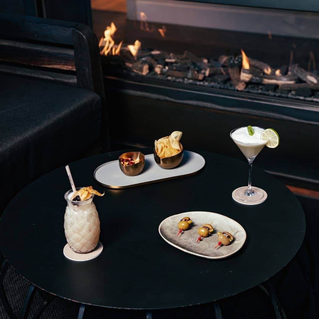 Two cocktails, chips, nuts, and olives are arranged on a black table by a modern fireplace&mdash;an inviting scene at one of the Best Restaurants in Barcelona.