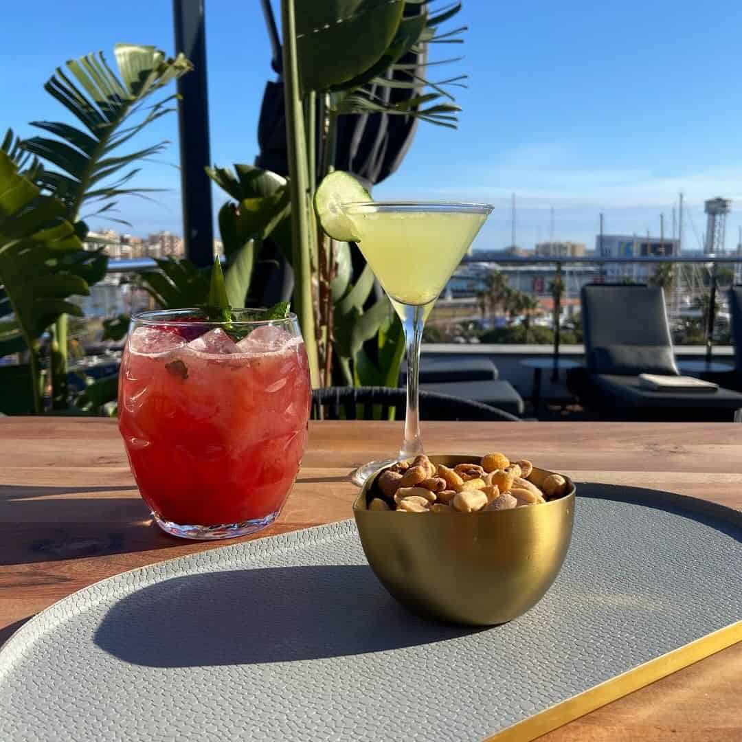 Two cocktails and a bowl of mixed nuts on a table, with a sunny outdoor lounge and marina in the background&mdash;perfect for those seeking Barcelona restaurants with a view.