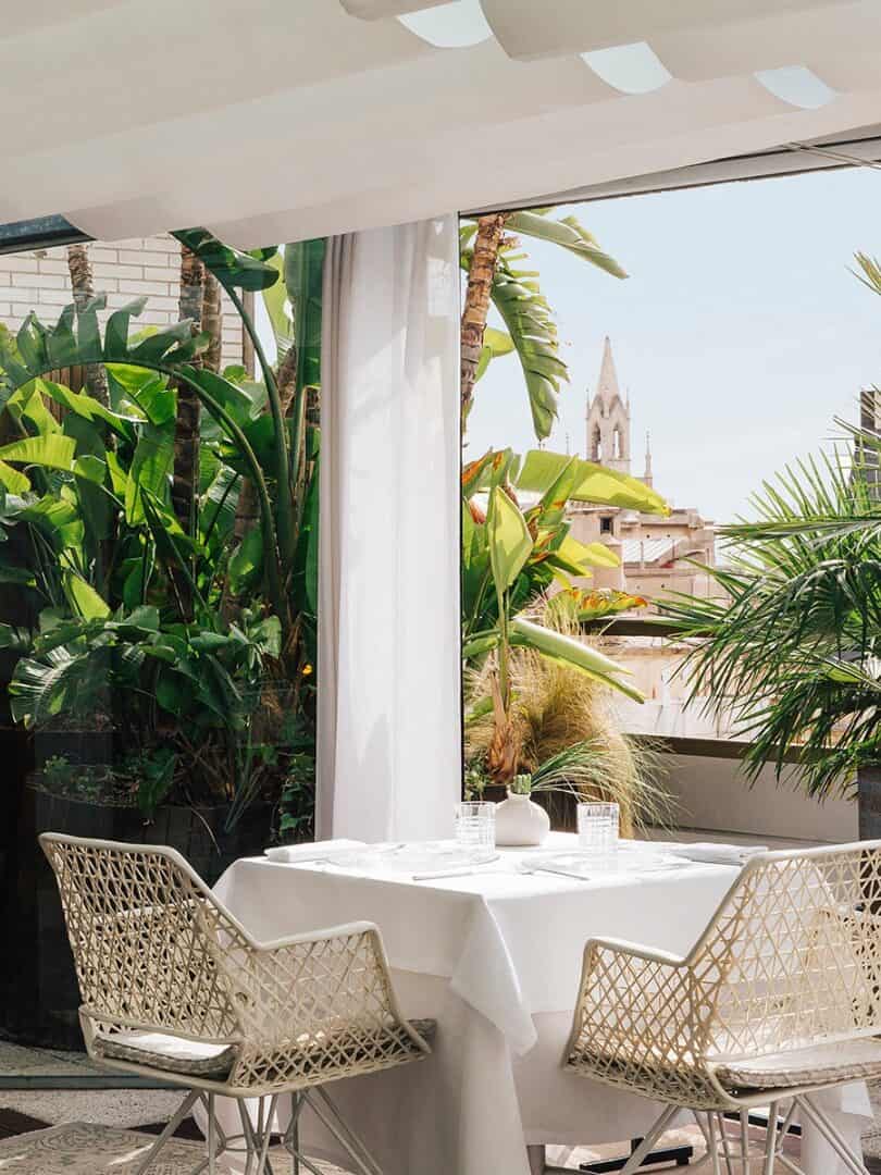 Outdoor dining area with a white table, chairs, lush green plants, and city buildings in the background&mdash;perfect for those seeking Barcelona restaurants with a view.