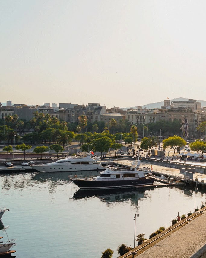 Yachts docked in a sunny Barcelona marina with city buildings and green trees in the background, near lively beachfront restaurants.