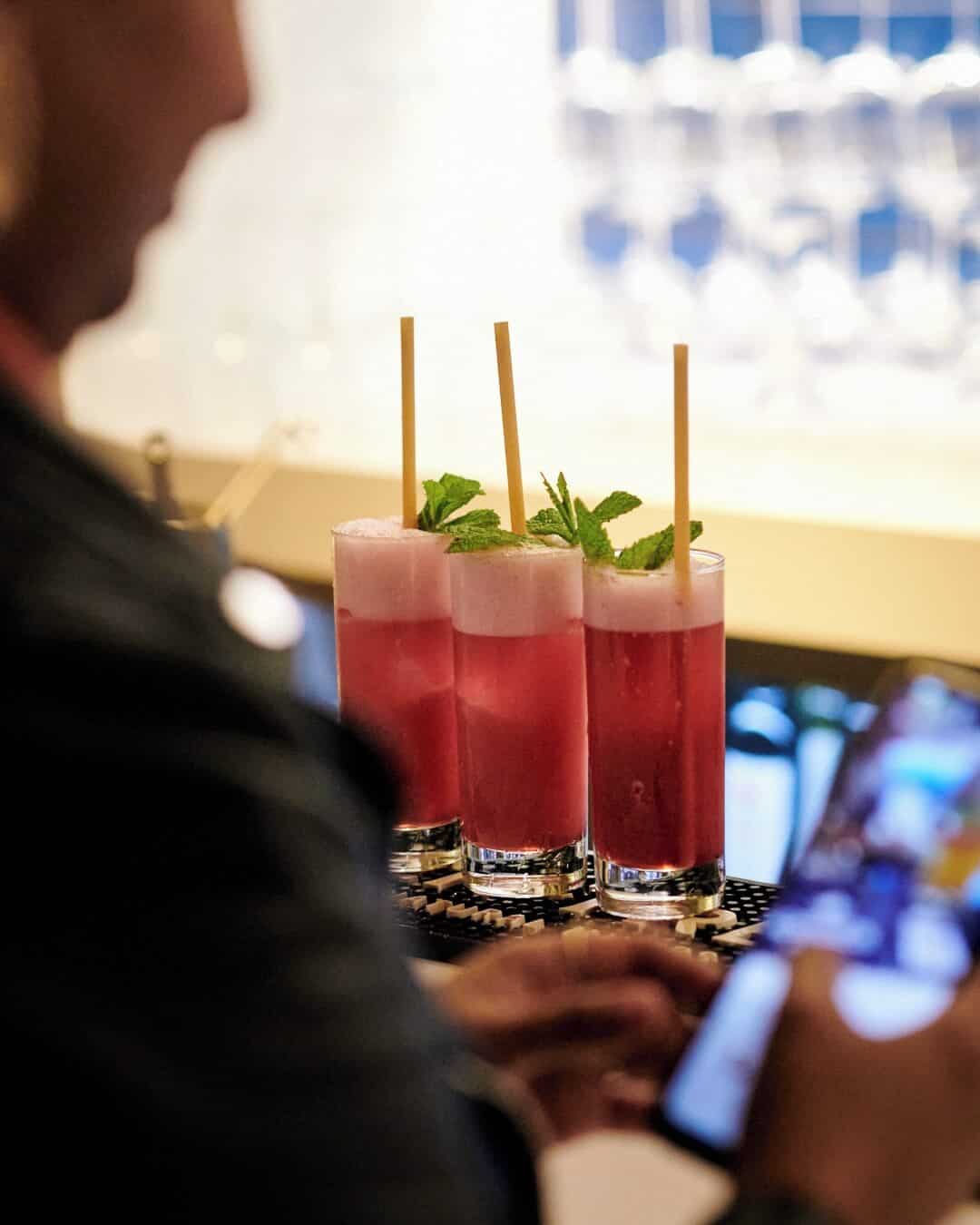 Three pink cocktails with mint garnish and straws sit on a bar, while a person checks their phone&mdash;perfect for capturing moments at one of the Best Restaurants in Barcelona.