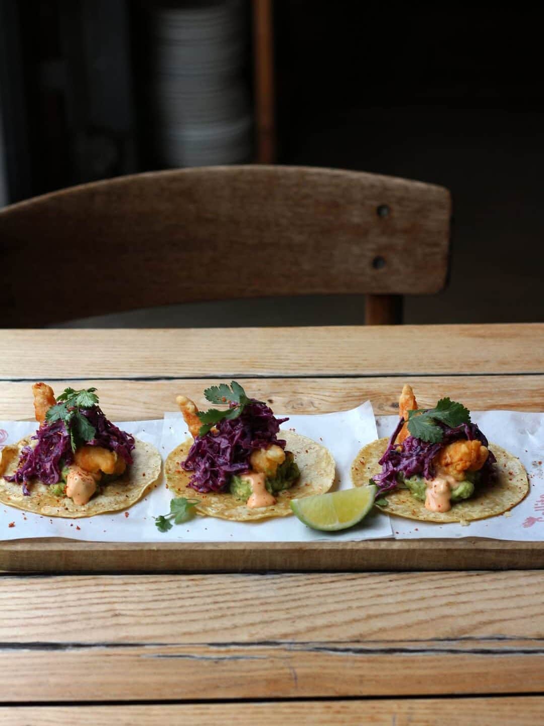 Three fish tacos topped with purple slaw and cilantro on a wooden table, with a lime wedge on the side&mdash;a flavorful taste of Barcelona dining.