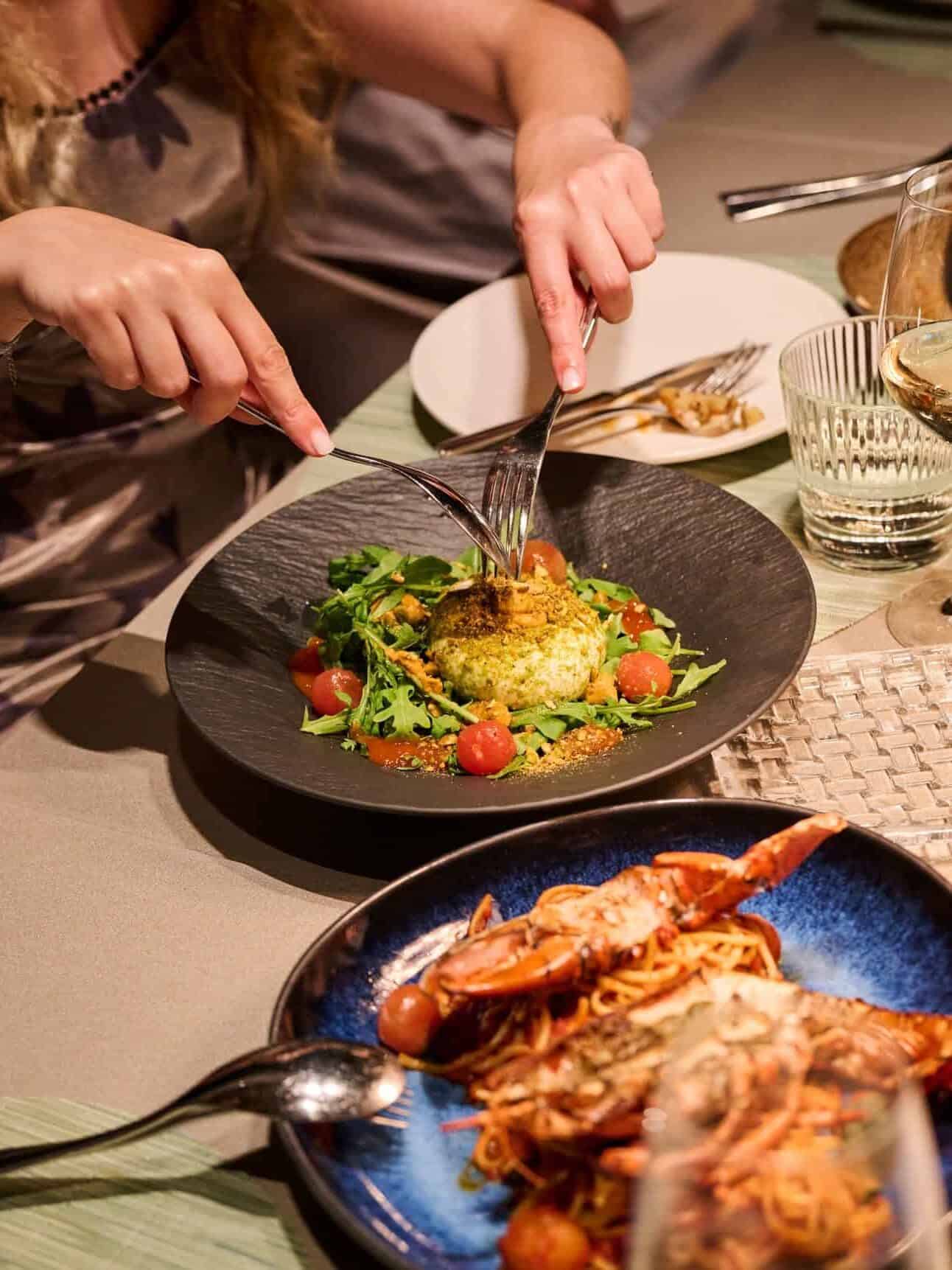 A person cuts into a salad with cherry tomatoes, arugula, and cheese; seafood pasta is nearby on the table&mdash;a scene you'd find in the Best Restaurants in Barcelona.