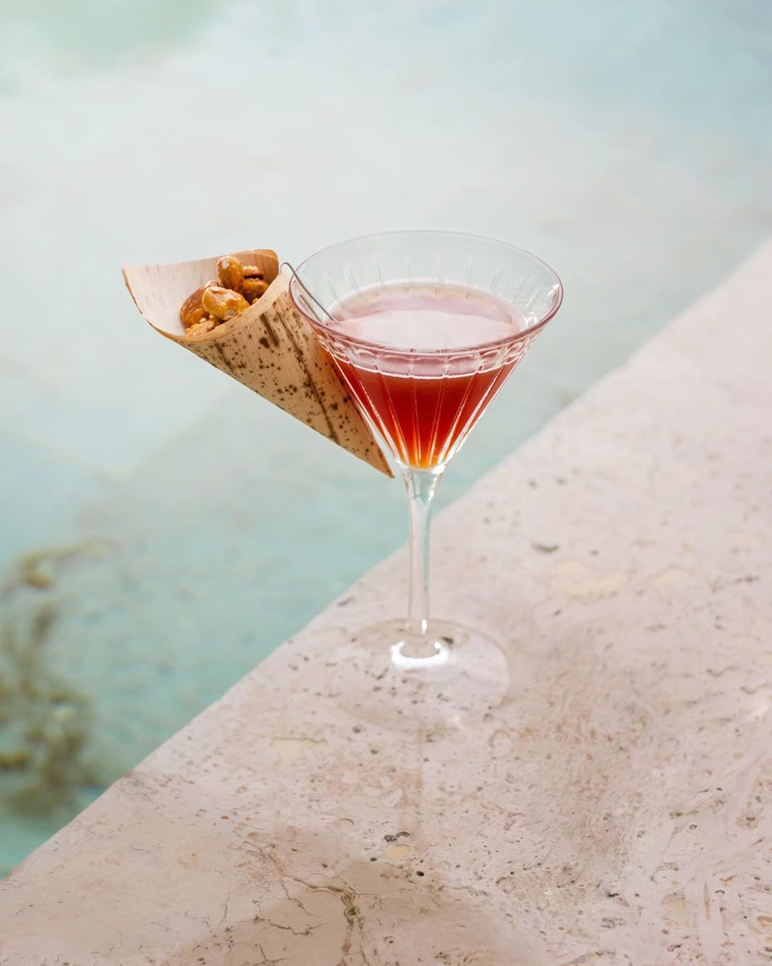 A pink cocktail in a martini glass with a cone of snacks, placed by the edge of a pool&mdash;perfect for relaxing at one of the best restaurants in Barcelona.