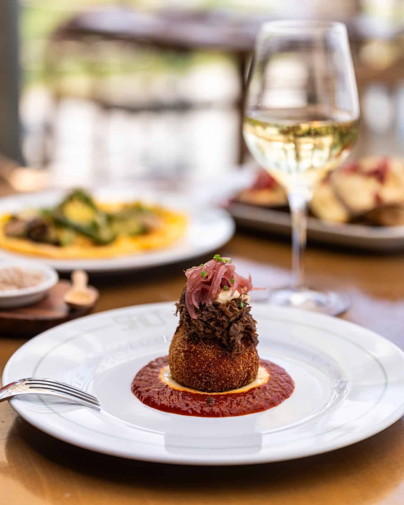 A gourmet dish with shredded meat on a fried base over red sauce, served with a glass of white wine—a perfect choice for those seeking Barcelona dining at its finest.