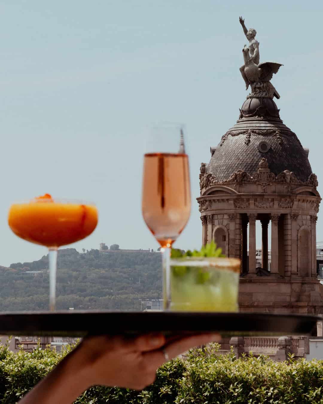 A tray with cocktails in front of a domed building and statue, with hills in the background&mdash;perfect for discovering restaurants with a view featured in any top Barcelona guide.