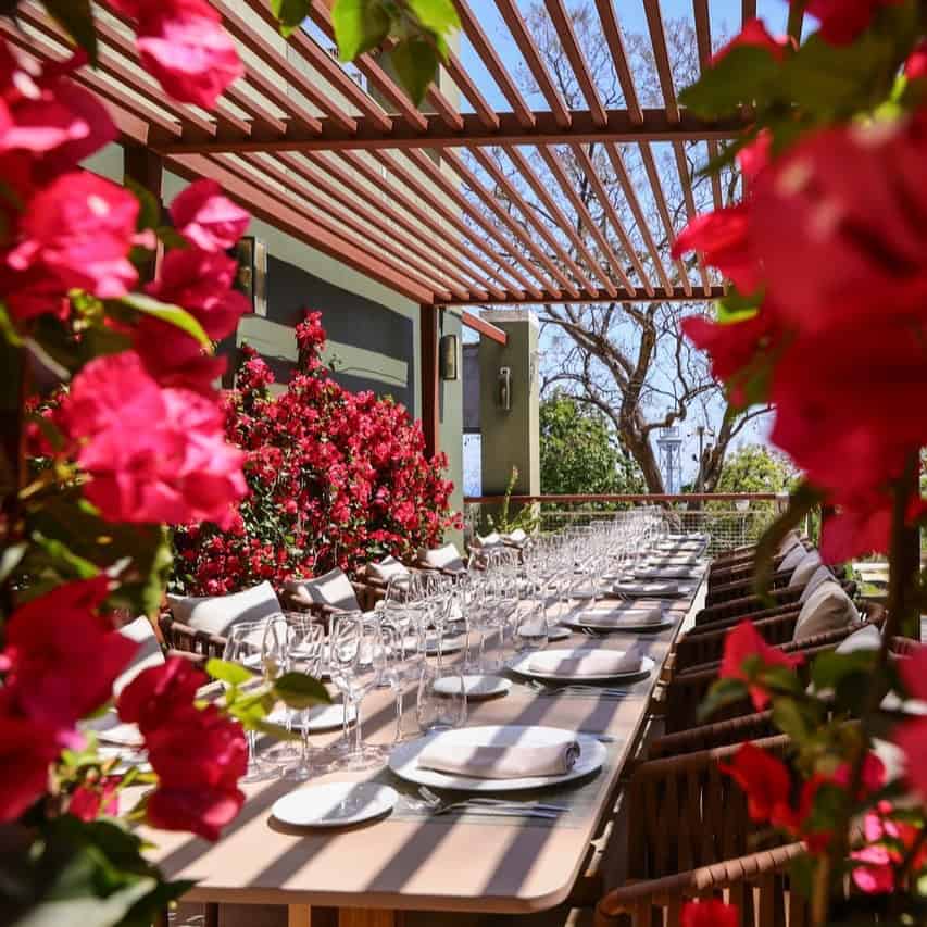 A long outdoor dining table set with plates and glasses, framed by vibrant pink flowers and greenery, evokes the relaxed charm of Barcelona dining.