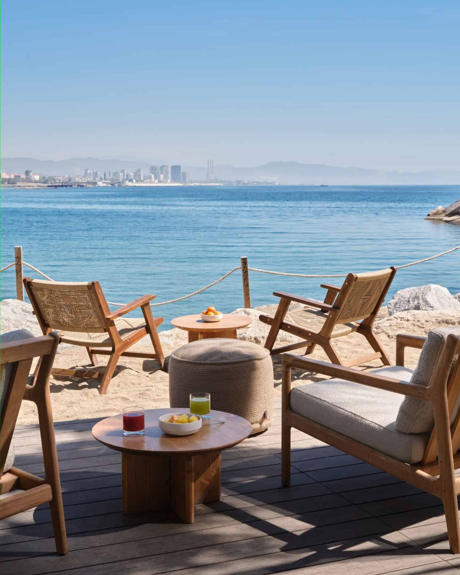 Wooden chairs and tables on a deck overlook a calm blue sea with a city skyline in the distance, capturing the relaxing ambiance of the best beachfront restaurants Barcelona has to offer.