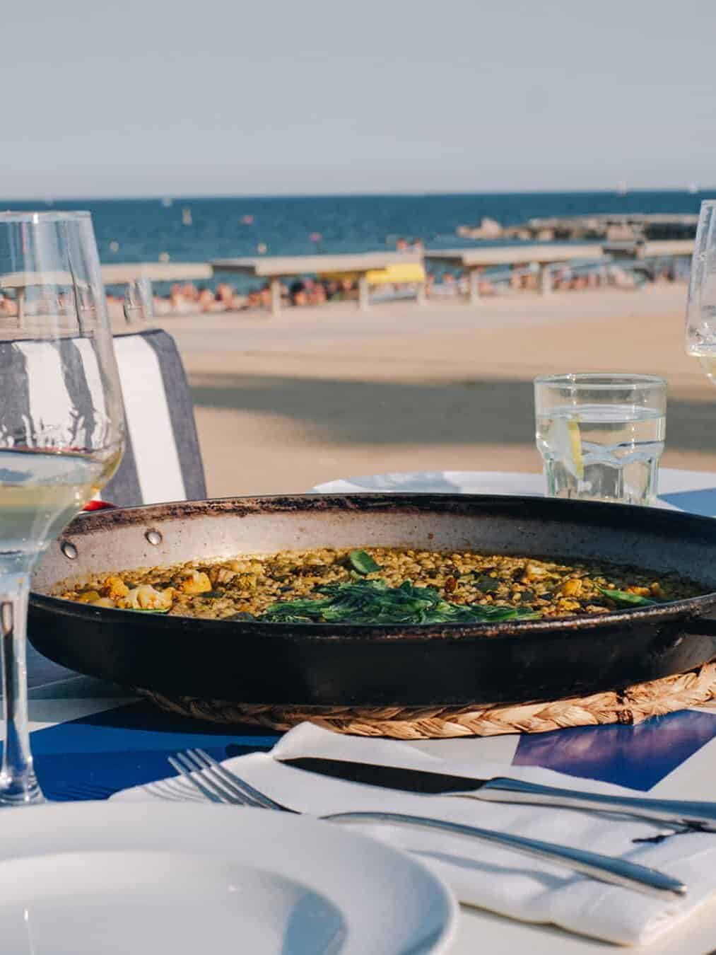 A pan of paella on a table by the beach at one of Barcelona’s vibrant beachfront restaurants, with wine glasses and the sea in the background.