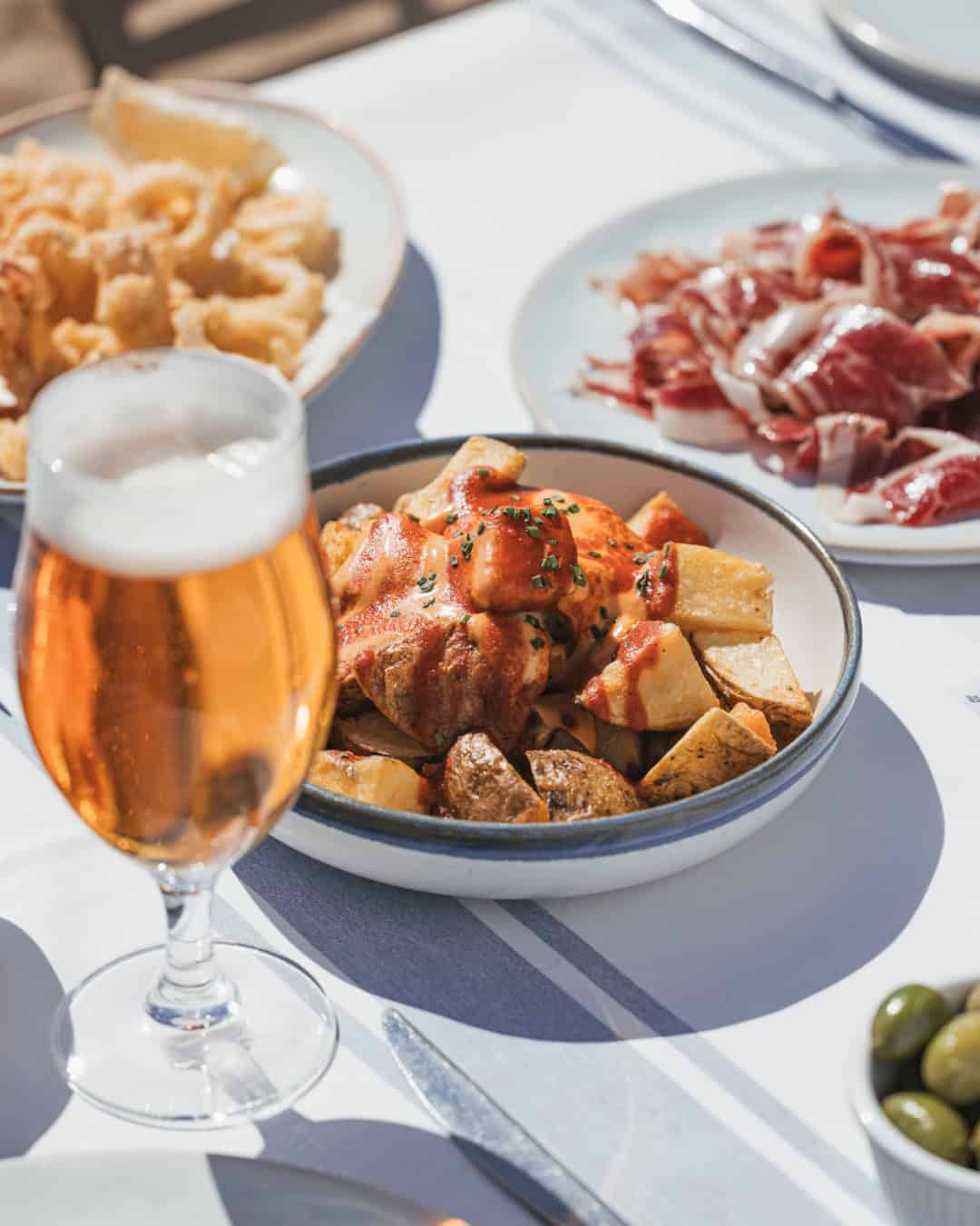 A glass of beer with plates of patatas bravas, fried calamari, and sliced cured ham on a table at one of Barcelona’s best restaurants.