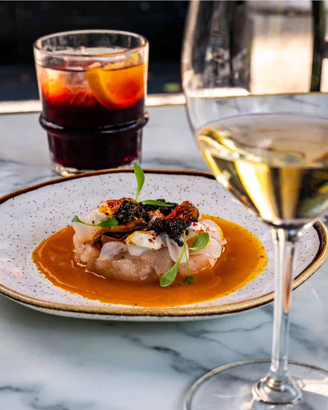 Savor a plated seafood dish with sauce, garnished herbs, white wine, and a cocktail on marble&mdash;an experience fit for the Best Restaurants in Barcelona.
