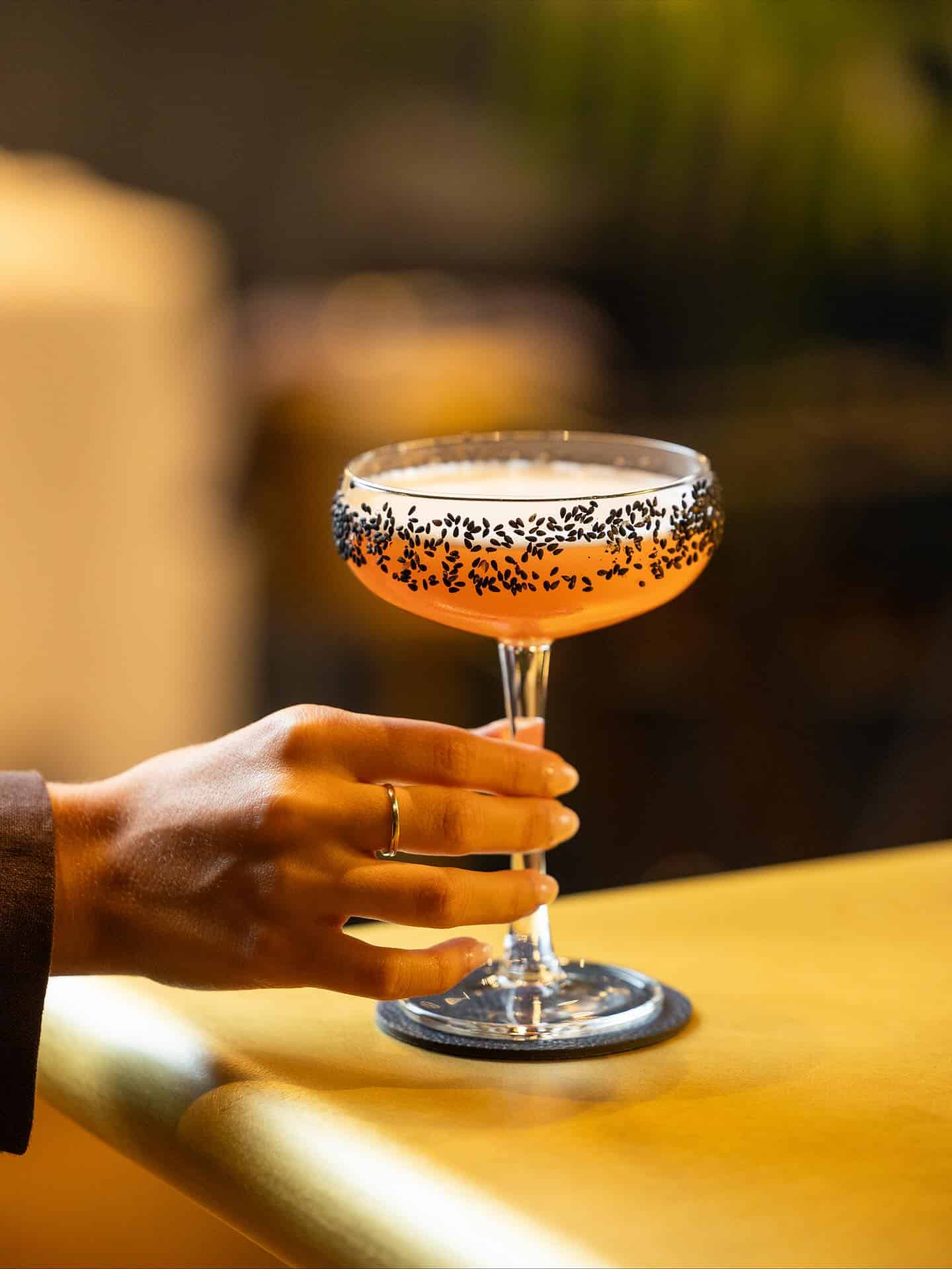 A hand holds a pink cocktail in a coupe glass with a black-sesame seed rim on a yellow bar&mdash;perfect for those searching for the Best Restaurants Barcelona has to offer.