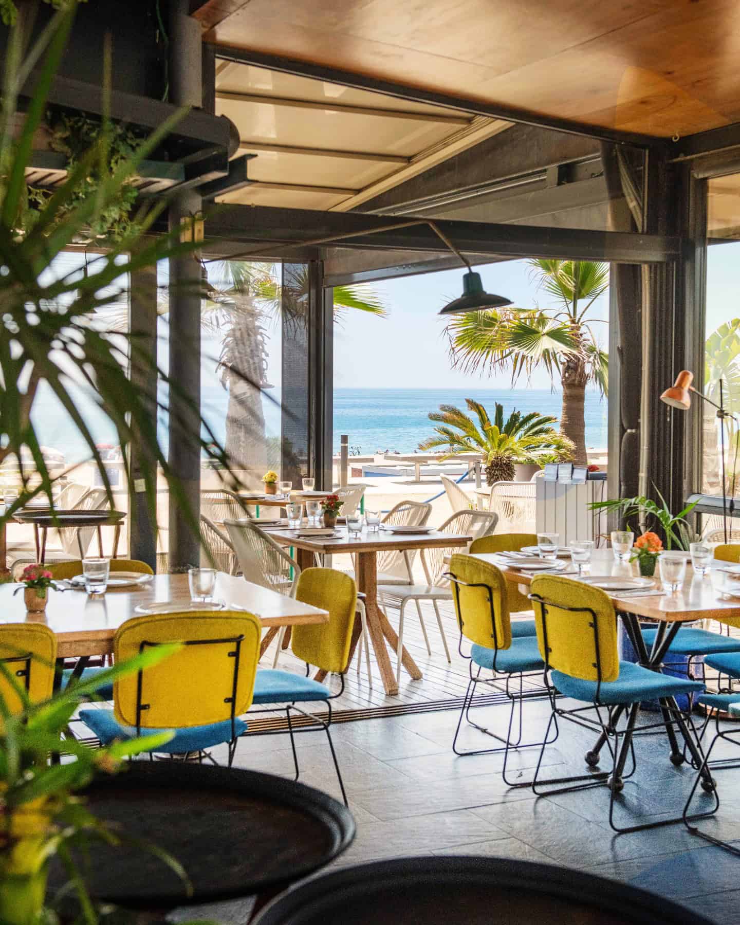 Bright seaside restaurant with yellow chairs, wooden tables, and a view of palm trees and the ocean&mdash;one of the best restaurants in Barcelona for dining with a view.