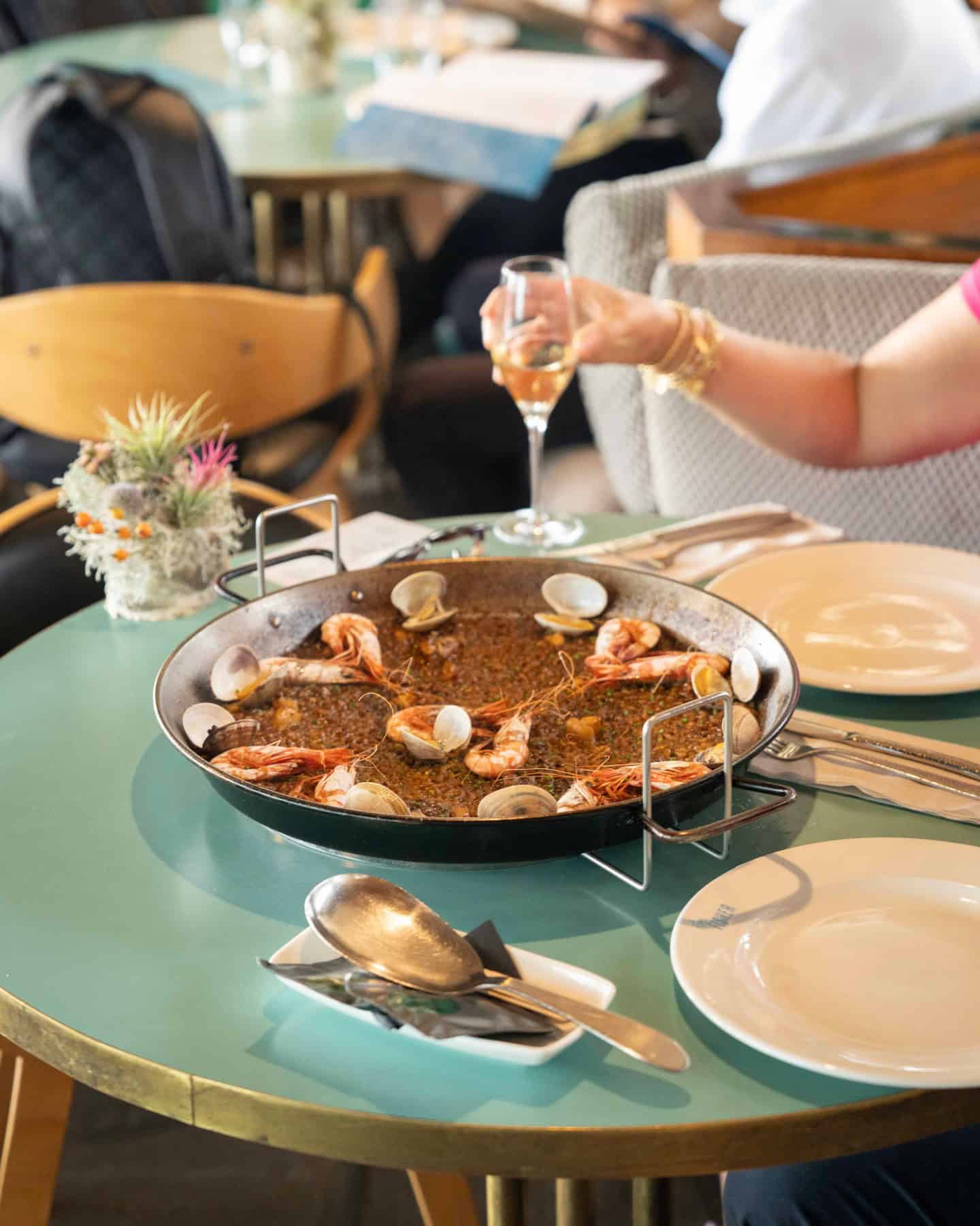 A pan of seafood paella sits on a table with plates, cutlery, and a person holding a glass of white wine&mdash;an ideal scene featured in any Barcelona Guide to the Best Restaurants in Barcelona.