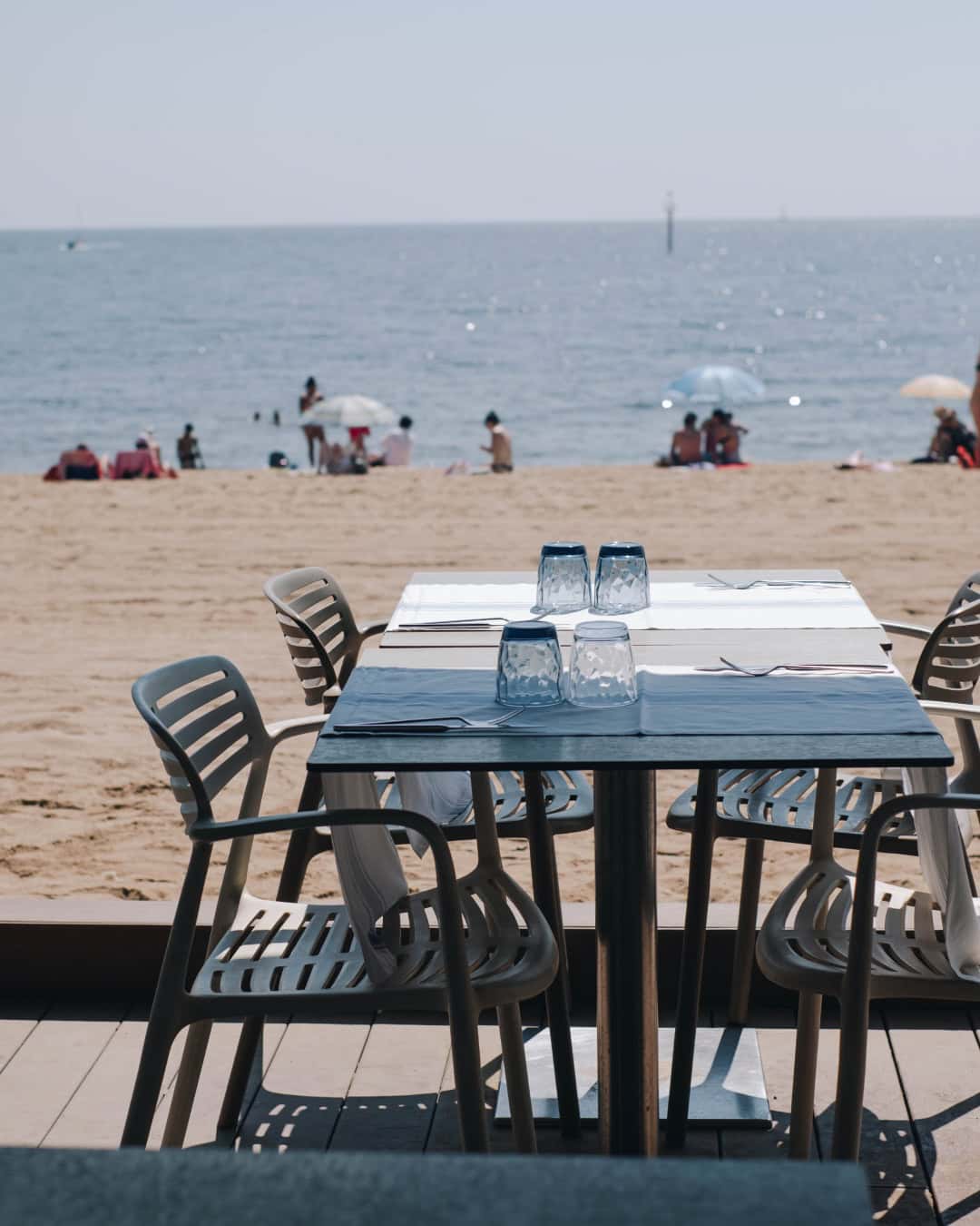 Outdoor dining table set for four overlooking a sandy beach with people and umbrellas by the sea—perfect for enjoying meals at one of Barcelona’s iconic beachfront restaurants.