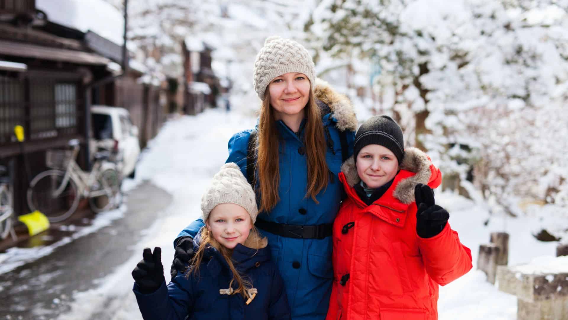A woman and two children in winter coats pose and smile on a snowy street, making peace signs.