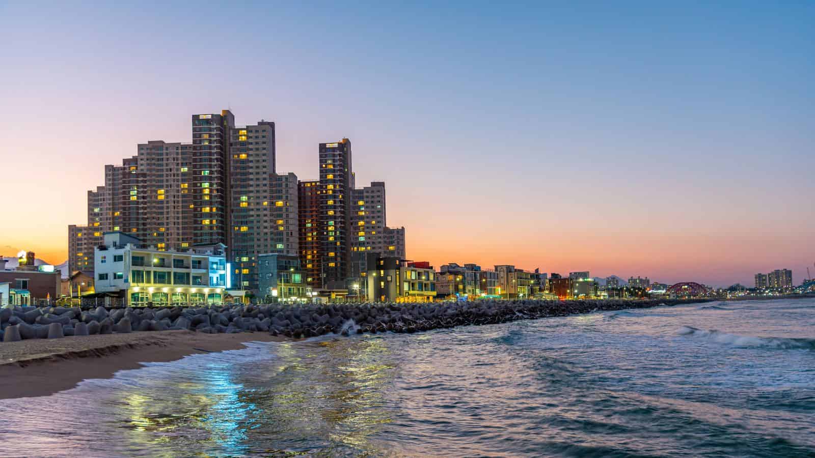 High-rise buildings and shoreline at sunset, city lights reflecting on calm ocean waves.