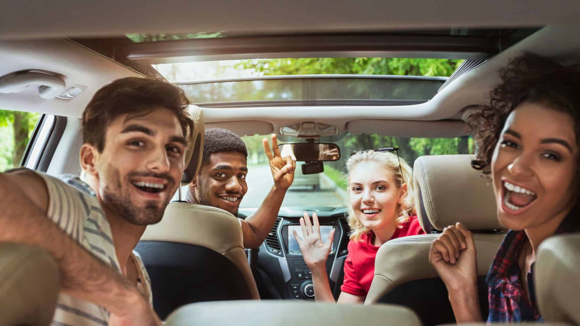 Four friends sitting in a car, smiling and waving at the camera, enjoying a road trip together.