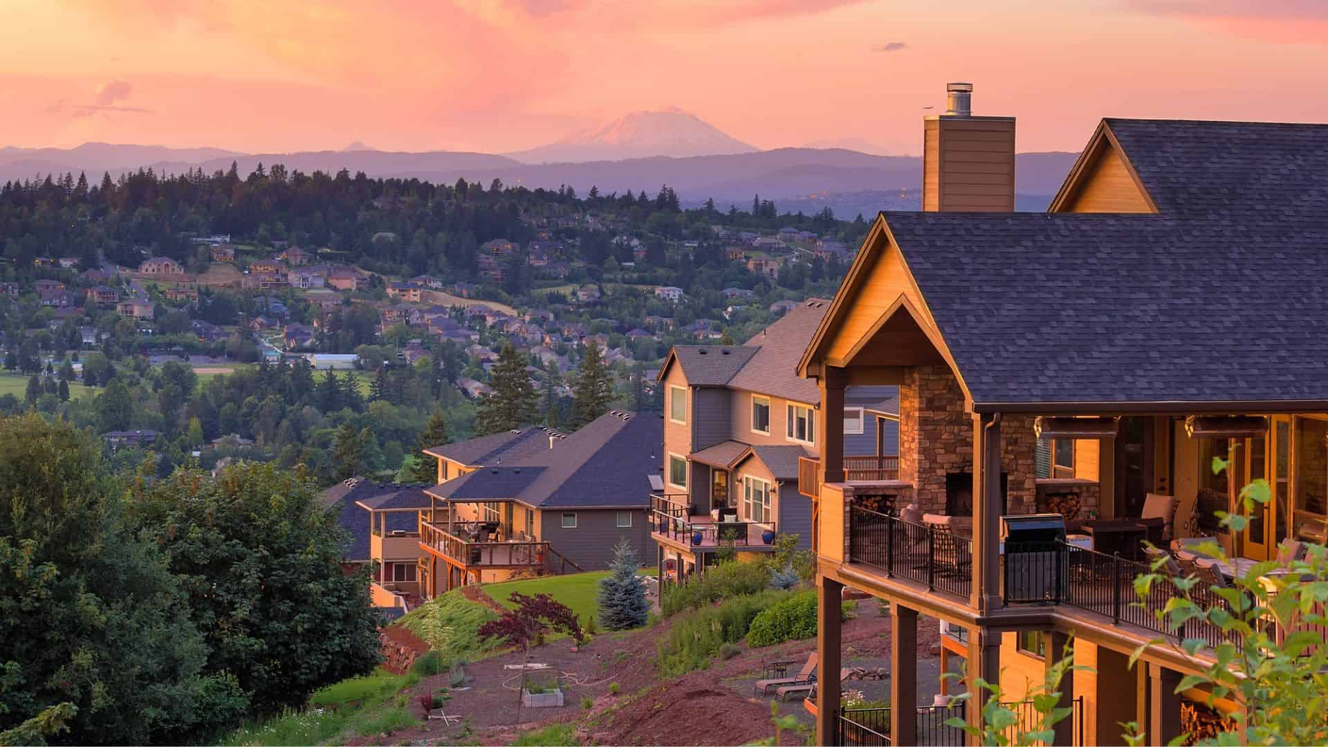 Large suburban houses on a hillside overlook a lush valley at sunset, with a distant mountain in the background.