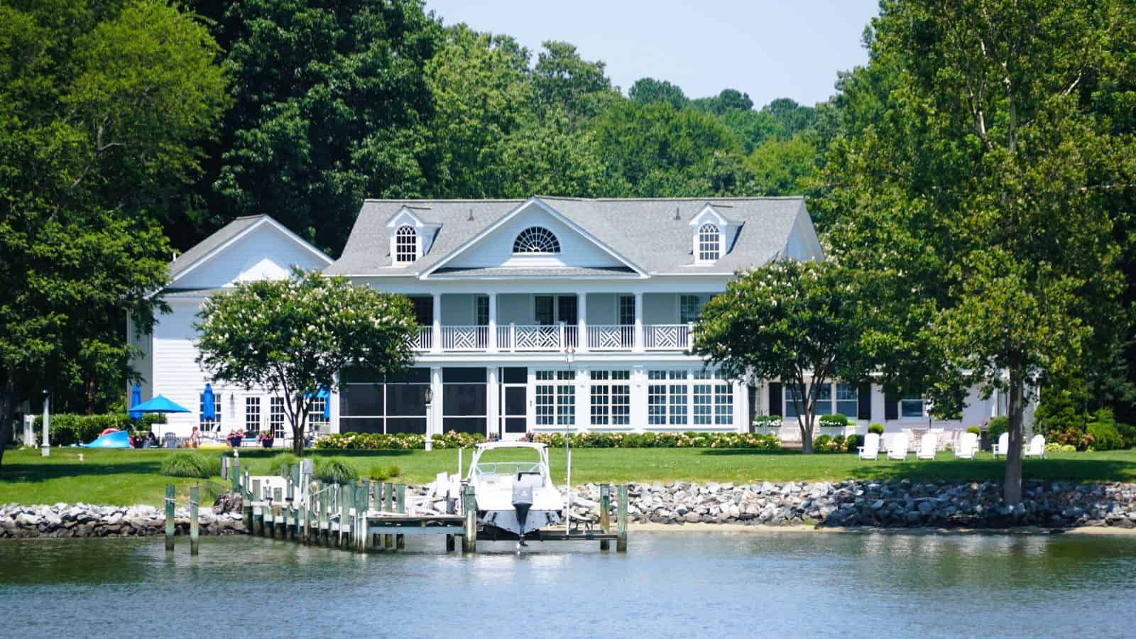 Large white lakeside house with boat dock, green lawn, and trees in the background.