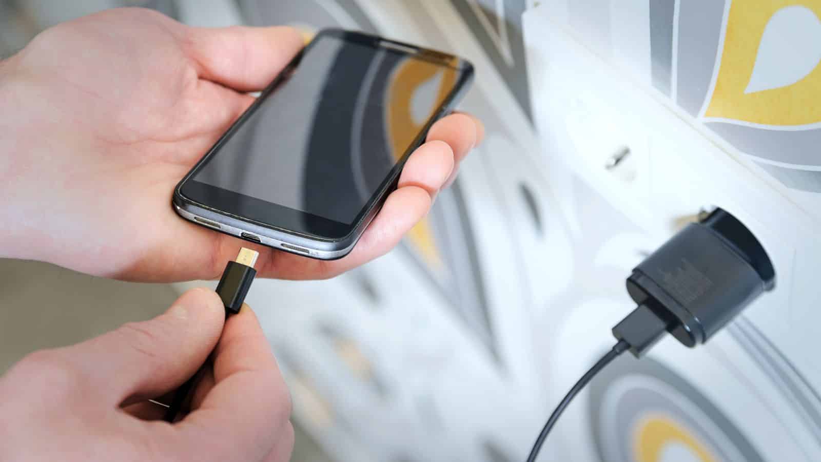 A person plugs a charging cable into a smartphone near a wall outlet with a plugged-in charger.