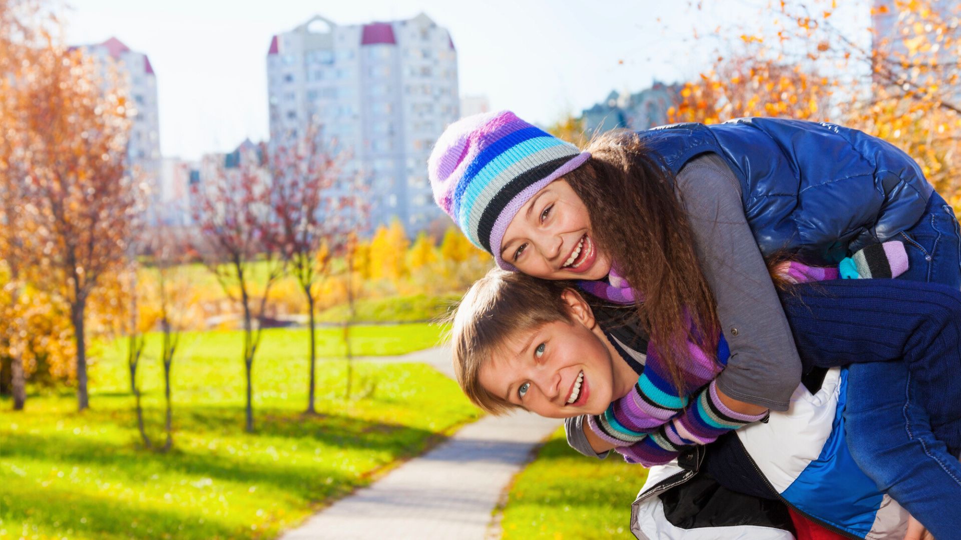 Two smiling children in winter clothes playing piggyback in a sunny park with autumn trees and buildings in background.
