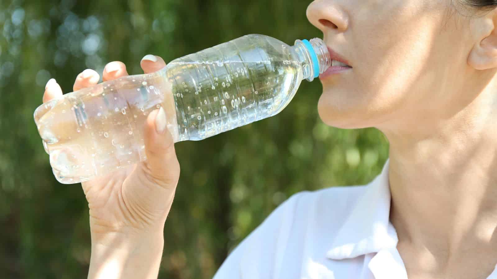 A woman holding and drinking from a clear plastic water bottle outdoors.