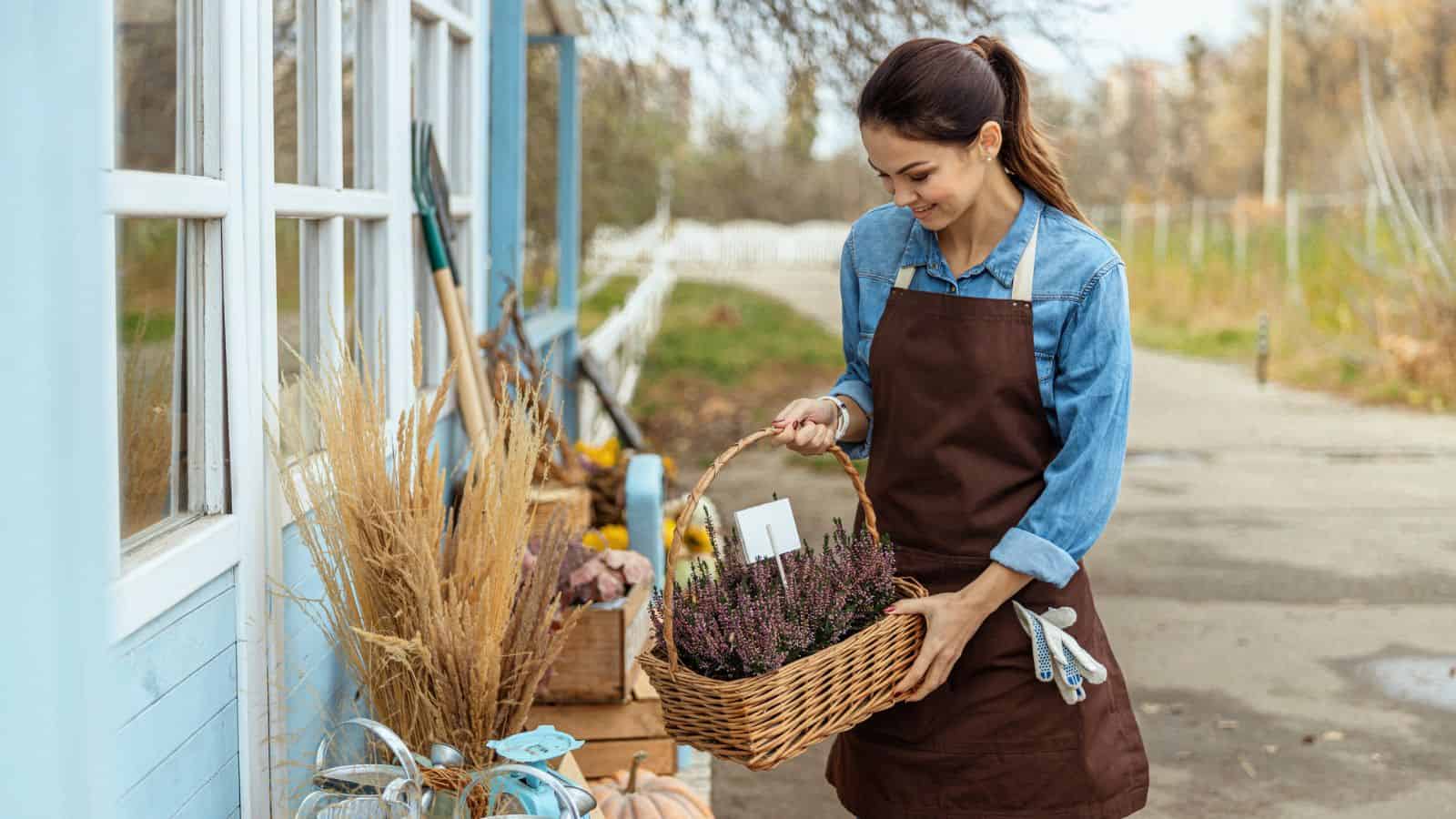 Woman in apron holding a basket of flowers outside a shop with gardening tools and plants on display.