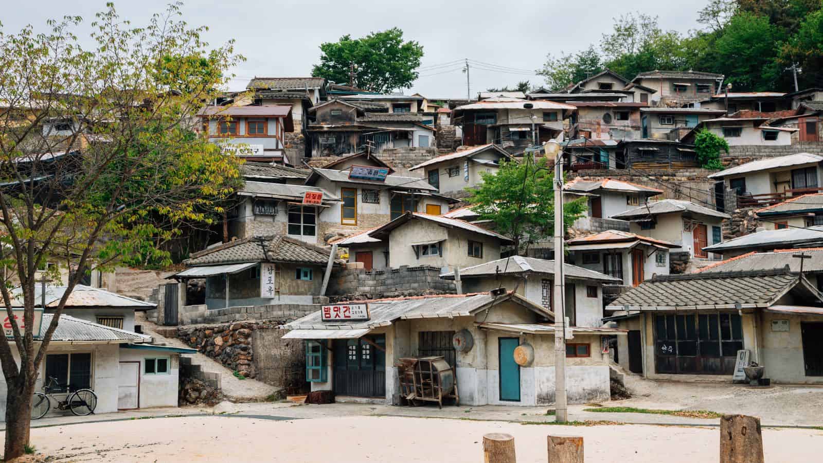 Cluster of small, old houses on a hillside with trees and a few signs in an outdoor setting.