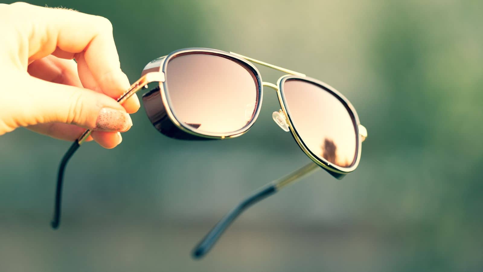 A hand holding aviator sunglasses with a blurred green background.