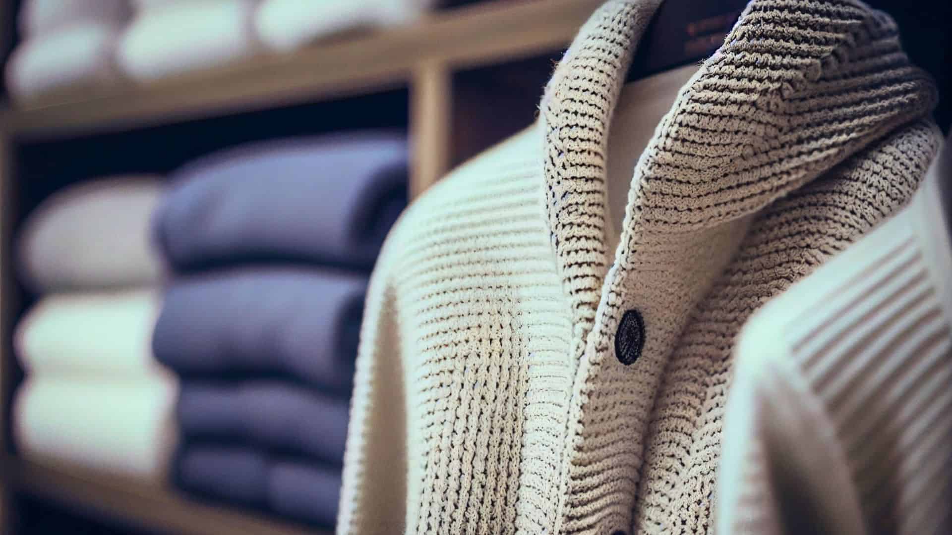 Close-up of a beige knit cardigan on a mannequin with folded sweaters on shelves in the background.