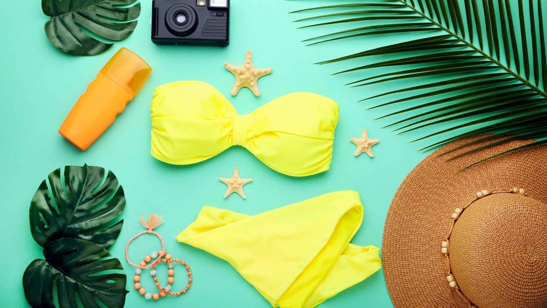 Flat lay of yellow bikini, straw hat, camera, sunscreen, jewelry, starfish, and tropical leaves on green background.