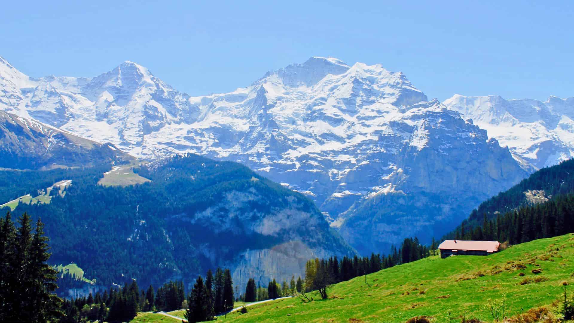 Snow-capped mountains with green hills, pine trees, and a small house under a clear blue sky.